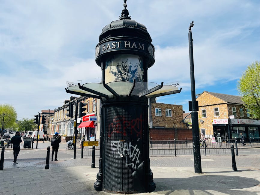 A cylindrical, black-and-white vintage-style street clock with a domed top and decorative finial, situated at a busy urban intersection in South Tottenham. The clock's face displays the time and the w