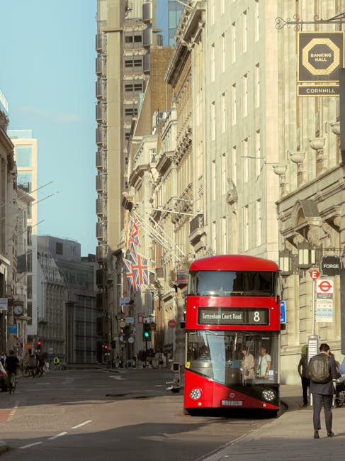A busy urban street scene featuring a red double-decker bus numbered 8 traveling along a paved road lined with historic beige and light gray buildings. The buildings display ornate architectural detai
